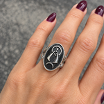 A close-up of a hand with dark purple nail polish wearing a silver ring featuring a black oval with a caridad del cobre design.