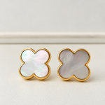 A pair of gold-tone stud earrings with four-leaf clover shapes set with iridescent mother-of-pearl inlays, displayed on a light beige surface.