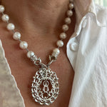 A sterling silver Caridad del Cobre pendant worn on a pearl necklace, displayed on a person's neck.
